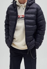 Hombre con una chaqueta de plumas negra sobre una sudadera crema con texto gráfico rojo y negro, combinado con pantalones negros que tienen un accesorio de cadena.