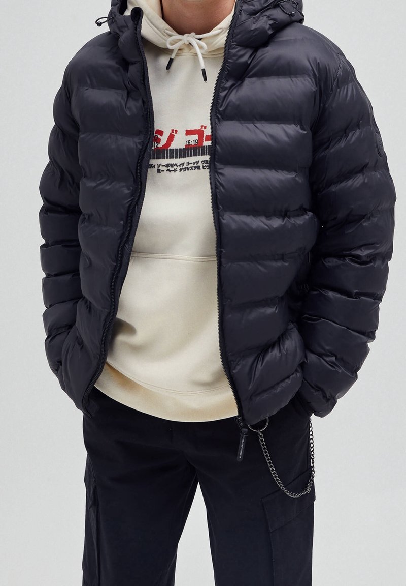 Hombre con una chaqueta de plumas negra sobre una sudadera crema con texto gráfico rojo y negro, combinado con pantalones negros que tienen un accesorio de cadena.