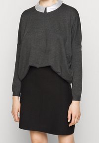 Kobieta ubrana w ciemnoszary sweter z długim rękawem z jasnoszarym kołnierzem oraz czarną spódnicę do kolan, stojąca na tle jednolitego tła.