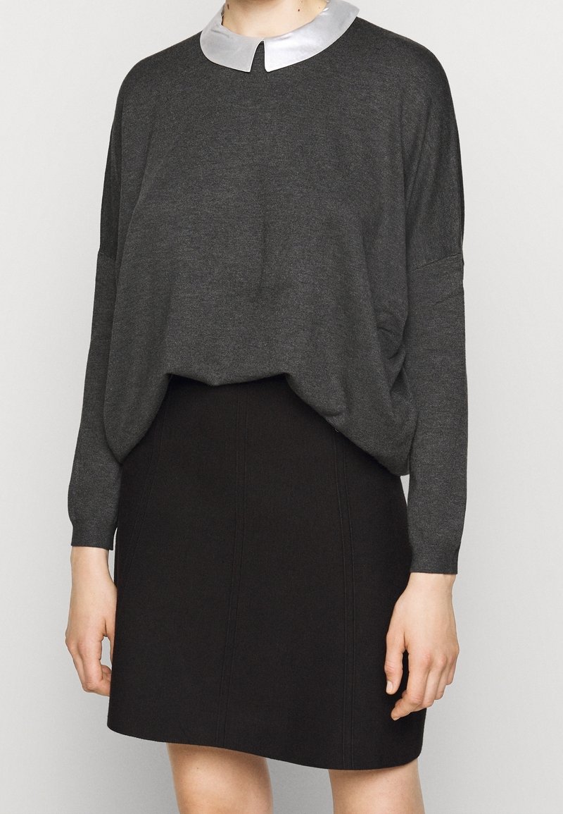 Kobieta ubrana w ciemnoszary sweter z długim rękawem z jasnoszarym kołnierzem oraz czarną spódnicę do kolan, stojąca na tle jednolitego tła.