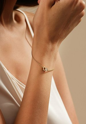 Goldarmband mit einer zarten Kette, die herzförmige Akzente und einen kleinen klaren Edelstein aufweist. Glatte Textur, minimalistisches Design.