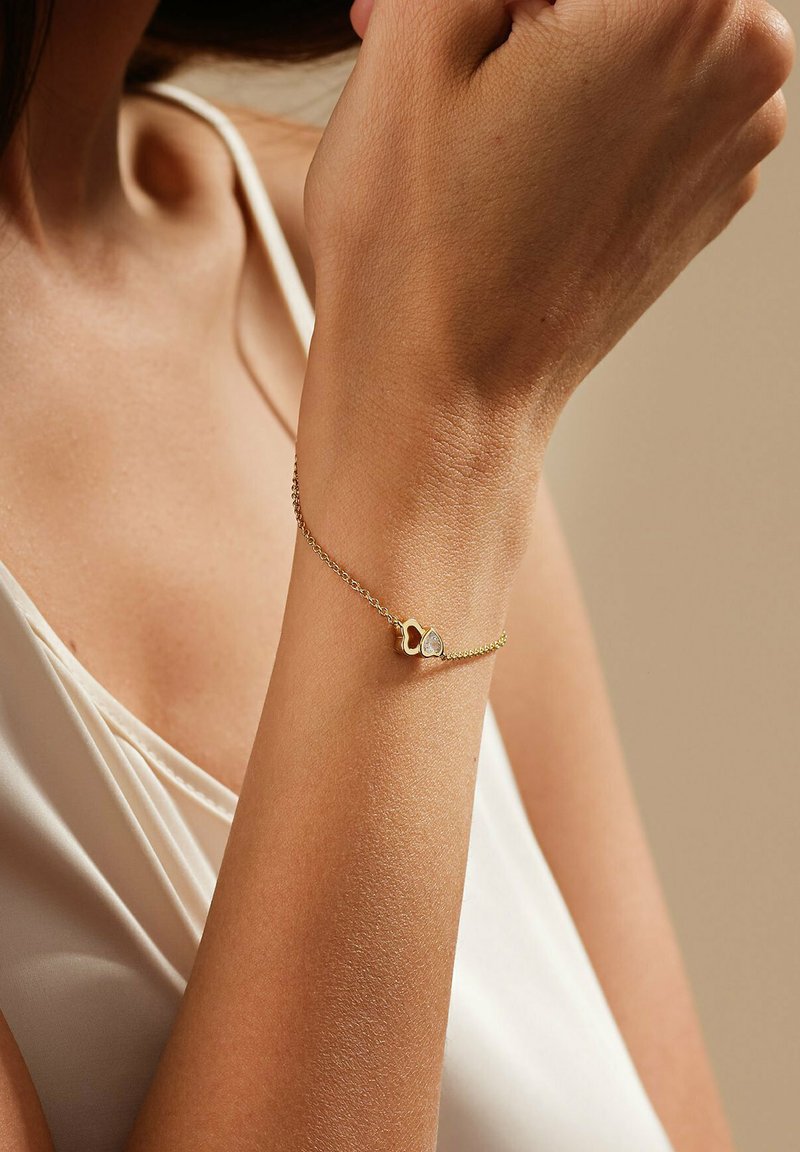 Goldarmband mit einer zarten Kette, die herzförmige Akzente und einen kleinen klaren Edelstein aufweist. Glatte Textur, minimalistisches Design.