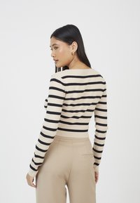 Sweter w paski w odcieniach beżu i czerni, dopasowany krój, ściągacze na mankietach i u dołu, teksturowany splot, noszony z beżowymi spodniami o szerokich nogawkach.