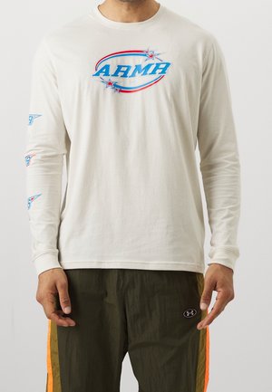 Homme portant une chemise blanche à manches longues avec le texte "ARMY" en bleu et rouge et des étoiles, et un pantalon vert foncé avec des bandes orange.