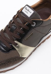 Brązowo-złoty sneaker Pepe Jeans z teksturowaną tkaniną, brązowymi sznurowadłami, czarnymi akcentami oraz oznaczoną metką na języku i boku.