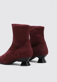 Botines de ante burdeos con un tacón negro brillante y redondeado, y un diseño elegante. Textura suave con detalles de costura a lo largo de los bordes.