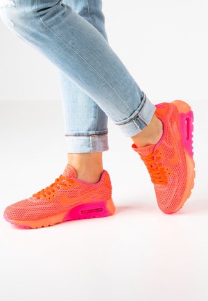 Person wearing light blue rolled-up jeans and bright orange-pink Nike sneakers, standing with one foot crossed behind the other.
