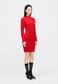 Rdeča rebrasta obleka z dolgimi rokavi in visokim ovratnikom, prilegajoča silhueta. Oblikovana z črnimi visokimi petami in prozornimi dokoleni. 