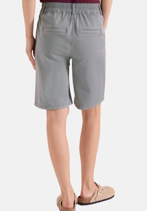 Persona con pantalones cortos grises hasta la rodilla con cinturilla elástica y bolsillos con tapeta en la parte trasera, combinado con sandalias beige sin cierre.
