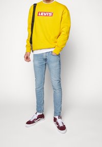 Gelbes Sweatshirt mit rotem Levi's-Logo, hellblaue Skinny Jeans und kastanienbraune Sneaker mit weißen Akzenten. Inklusive schwarzem Schultergurt.