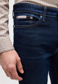Tamno plave traperice s žutim šavovima, s Calvin Klein oznakom na stražnjem džepu. Ruka počiva na bedru.