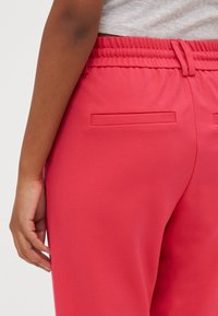 Detailní záběr osoby v červených elastických kalhotách s kapsami na zadní straně a světle šedém topu, ruka uvolněná podél těla.