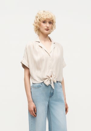 Mladá žena s kučeravými blond vlasmi, ktorá má na sebe béžovú pruhovanú krátku blúzku zviazanú v páse a svetlomodré rifle s vysokým pásom.