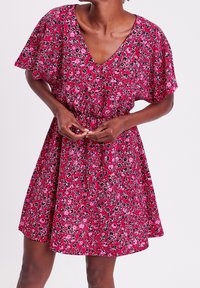 Floral short dress in pink with black and purple patterns. V-neck, short sleeves, and an elastic waistband for a relaxed fit. Lightweight fabric.