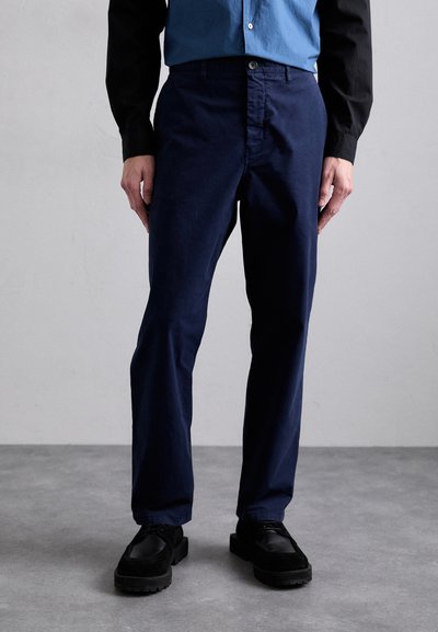 Pantalon bleu marine à coupe droite, avec fermeture à bouton et poches latérales, fabriqué dans un tissu lisse, porté avec des chaussures noires.
