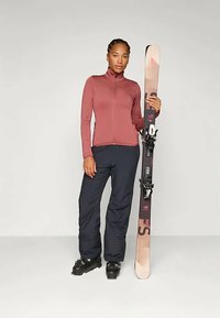 Síruha, amely egy testhezálló, bordó cipzáras dzsekit és tengerészkék nadrágot tartalmaz. Az illető egy sílécet tart kötéssel, valamint fekete sícipőt visel.