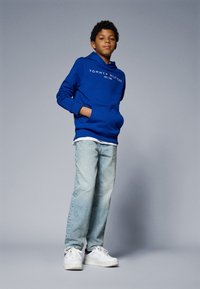 Blå bomullshoodie med vit logotyp, bärs över en vit skjorta, i kombination med ljusblå denimjeans och vita sneakers.