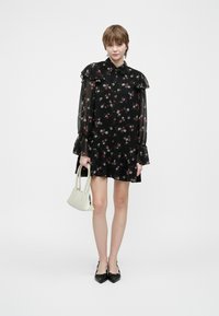 Γυναίκα φορώντας ένα μαύρο floral φόρεμα με μακρύ μανίκι, κρατώντας μια μικρή λευκή τσάντα και φορώντας μαύρα παπούτσια με μυτερή μύτη, μπροστά σε μία απλή φόντο.