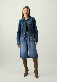 Chaqueta de mezclilla en azul oscuro, abotonada con bolsillos en el pecho; combinada con pantalones cortos de mezclilla de pierna ancha en azul claro y botas marrones hasta la rodilla.
