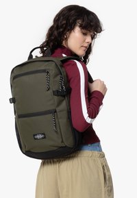 Sac à dos en tissu vert avec des accents noirs, plusieurs poches avant et une surface texturée. Comprend des sangles réglables et des poignées de transport renforcées.