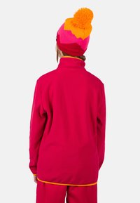 Klarrrosa fleecejacka med hög krage, med en gul accent vid fållen, parat med en livlig rosa och orange mössa med en pom-pom.