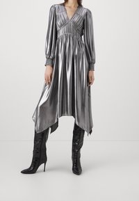 Robe argentée brillante avec de longues manches bouffantes, un décolleté en V et une taille froncée, dotée d'un ourlet asymétrique. Associée à des bottes noires à talons hauts ajustées.