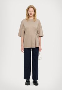 T-shirt loose-fit di colore taupe con maniche oversize e dettaglio a forma di anello in argento in vita, abbinato a pantaloni larghi blu navy e scarpe nere.