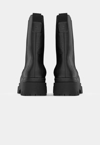 Par svart lærstøvler med klumpete såler, sett bakfra mot en ensfarget hvit bakgrunn.