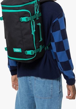 Persoon die een zwarte en turquoise Eastpak-rugzak draagt, gekleed in een blauw geruite trui en lichtblauwe jeans, van achteren getoond.