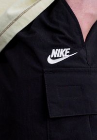 Čierna tkaninová košeľa s výrazným bielym logom Nike, s detailom vrecka na prednej strane a béžovým lemom na golieri.