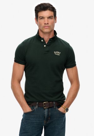 Donkergroen katoenen polohemd met een korte kraag, twee knopen bij de sluiting en een geborduurd logo. Draagt een blauwe spijkerbroek en een bruine riem.