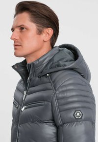 Giacca a vento grigia con cappuccio, caratterizzata da cuciture orizzontali, tasche con zip e un'etichetta patch a forma esagonale sulla spalla.