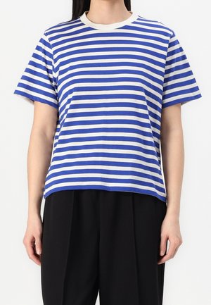 Personne portant une chemise à manches courtes rayée horizontalement bleu et blanc associée à un pantalon noir ample, debout devant un fond uni.