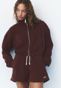 Woman wearing a dark brown zip-up sweatshirt and matching shorts with white drawstrings and "self." printed on the left leg.