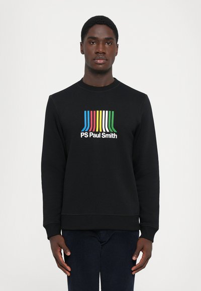 Mężczyzna w czarnej bluzie z kolorowym paskowanym wzorem i napisem "PS Paul Smith" stoi na białym tle.