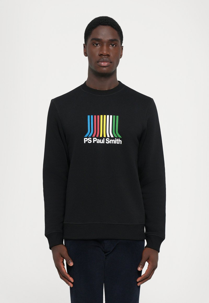 Mann iført svart hettegenser med fargerik stripete design og "PS Paul Smith"-tekst stående mot hvit bakgrunn.