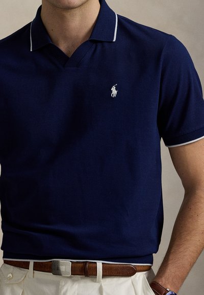 Μπλε ναυτικό πόλο μπλουζάκι με λευκή επένδυση στο γιακά και κεντημένο λογότυπο. Μαλακό ύφασμα, κοντά μανίκια και άνετη εφαρμογή. Φορεμένο με καφέ ζώνη.