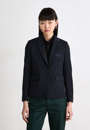 Blazer marine à revers, fermeture à un bouton, deux poches avant et détail brodé sur la poche poitrine gauche. Assorti à un pantalon vert.