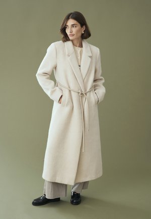 Beżowy wełniany płaszcz z paskiem w talii, szerokimi klapami i bocznymi kieszeniami, zestawiony z jasnym swetrem i spodniami w paski. Czarne buty.