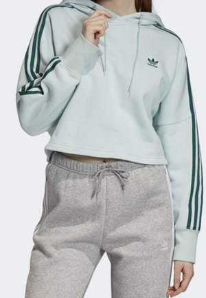 Haut à capuche cropped bleu clair avec un design à trois rayures vertes sur les manches et un logo, associé à un pantalon de survêtement gris avec des rayures blanches sur les côtés.