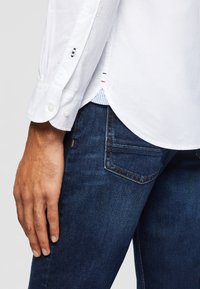 Gros plan d'une personne portant une chemise blanche boutonnée et un jean bleu foncé, montrant le poignet de la chemise et l'ourlet latéral sur le jean.