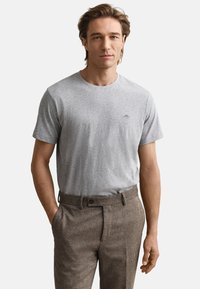 Siva pamučna majica s okruglim izrezom, uparena s smeđim teksturiranim hlačama. Majica ima mali izvezeni logo na prsima.