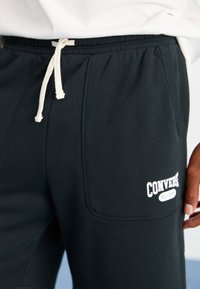 Black sweatpants with a drawstring waist, side pockets, and a white "Converse All-Star" logo on the thigh. Smooth fabric texture.