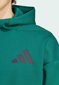 Zelena kapuća sa teksturiranim završetkom, sa crnim Adidas logotipom u trouglastom obliku na grudima. Mekani materijal i visoki ovratnik.