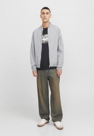 Chaqueta bomber gris claro con puños y cuello de canalé, llevada sobre una camiseta gráfica negra. Jeans oscuros de corte holgado en verde oscuro y zapatillas blancas.