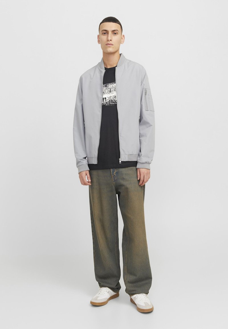 Chaqueta bomber gris claro con puños y cuello de canalé, llevada sobre una camiseta gráfica negra. Jeans oscuros de corte holgado en verde oscuro y zapatillas blancas.