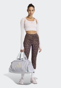 Ανοιχτό γκρι τσάντα duffel με λευκές λουριά και μεγάλο λογότυπο. Το μοντέλο φοράει ένα απαλό ροζ κοντό τοπ και καφέ κολάν με πλάτυ μοτίβο λεοπάρδαλης.