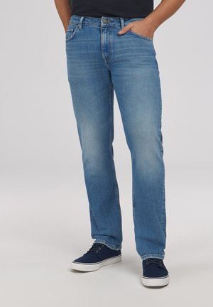 Mężczyzna w jasnoniebieskich prostych dżinsach z rękami w kieszeniach oraz granatowych trampkach z białymi podeszwami.