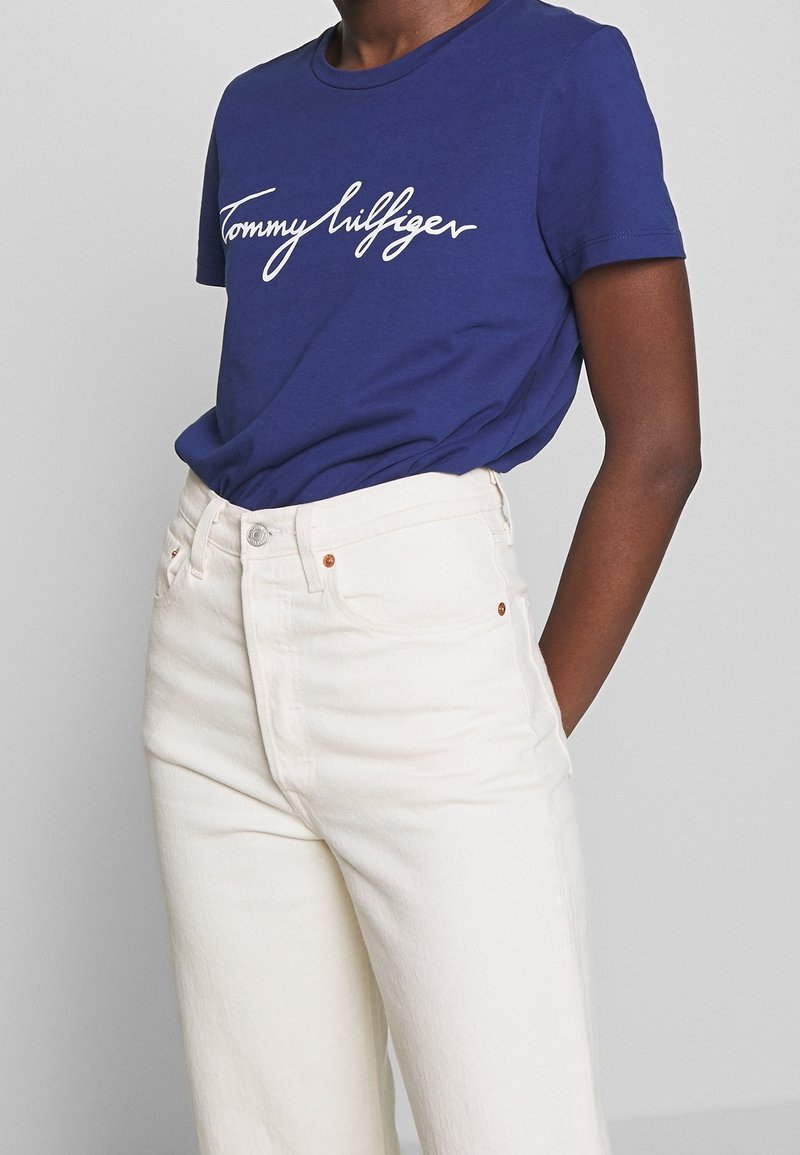 Modrá bavlněná tričko s bílým kurzívním logem "Tommy Hilfiger", spárované s krémovými džíny s vysokým pasem. Jednoduchý design s krátkým rukávem.