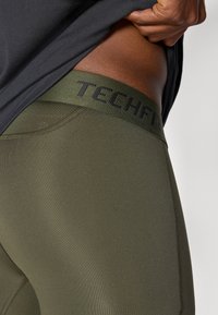 Primer plano de una persona levantando una camiseta negra para revelar la cinturilla de unas mallas deportivas TECHFIT de color verde oliva.